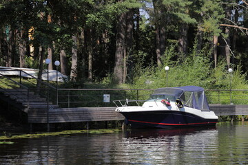 Beautiful view on parked modern cabin motor boat near Pier in forest marina with green trees on shore, river calm water at quiet summer day, boating recreation