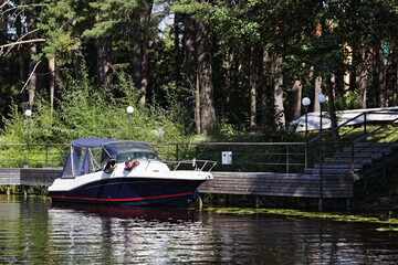 Fototapeta na wymiar One parked motor boat near Pier in forest bay marina with green trees on shore, river calm water at quiet Sunny summer day, boating recreation