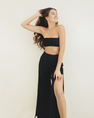 Fototapeta premium young brunette girl with long hair in black costume is standing on the white wall background with hand in hair and smiling happy