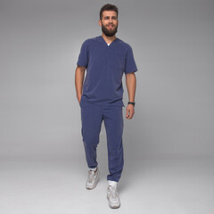 young stylish bearded male nurse with long legs in blue medical costume is standing in step with hand in pocket, looking straight and smiling cute on gray wall background. medical concept. free space