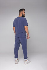Fototapeta premium young stylish bearded nurse male in blue medical costume is standing from the back and smiling cute on the white wall background. medical fashion concept. free space