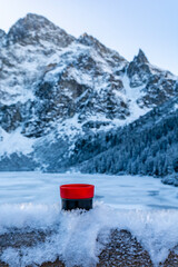 A red and black thermos cup with a hot drink (tea) placed in the frozen snow against the background of mountain peaks near the Morskie Oko Lake. Tatra Mountains, Poland.