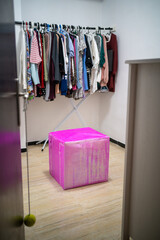 A giant square box wrapped with shiny pink gift wrap awaits to be opened in a lightened room used as a wardrobe