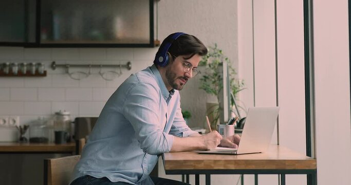 Young concentrated man student sit at home wear modern wireless headphones, listens audio lecture course on-line, writing important notes. Knowledge improvement, self-education using internet concept