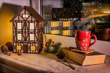 On windowsill there is half-timbered house nightlight and red Christmas mug among Christmas decorations against the background of snowy window at night.