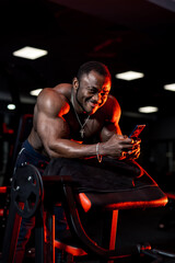 Fototapeta na wymiar African sportsman with perfect body smiles and reads message in gym. Dark light background.