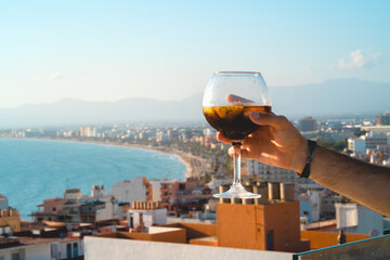A hand raises a cup of an alcoholic cocktail froma roof top bar in a hotel at Mallorca with the bay...