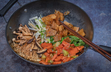 Vegan stir fry in black wok with cooking chopsticks on gray background, copy space