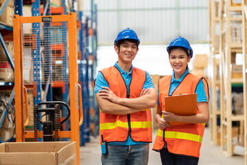 Portrait of smiling Asian male and female warehouse worker in safety vest and helmet standing with arms crossed, holding clipboard in storage warehouse with shelf pallet spare parts parcel background