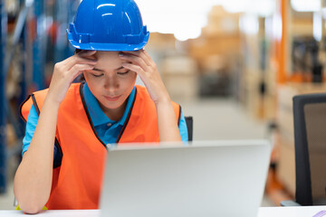 Asian female worker in safety vest and helmet sitting and working with computer laptop in storage warehouse. people, warehouse and industry concept
