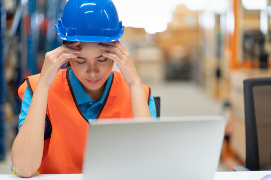 Asian Female Worker In Safety Vest And Helmet Sitting And Working With Computer Laptop In Storage Warehouse. People, Warehouse And Industry Concept