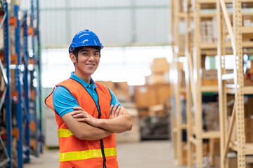 Confident smiling Asian male warehouse worker in safety vest and helmet standing with arms crossed...