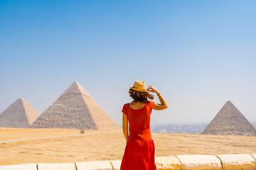 A tourist girl in a red dress looking at the Pyramids of Giza, the oldest Funerary monument in the world. In the city of Cairo, Egypt - obrazy, fototapety, plakaty