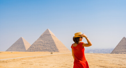 A tourist girl in a red dress looking at the Pyramids of Giza, the oldest Funerary monument in the...