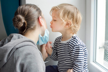 Child in home quarantine taking the medical mask off his mothers face and kissing her during coronavirus COVID-2019 and flu lock down. Selective focus