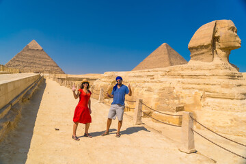 A tourist couple at The Great Sphinx of Giza and in the background the Pyramids of Giza, the oldest Funerary monument in the world. In the city of Cairo, Egypt - obrazy, fototapety, plakaty