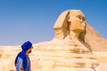 Fototapeta na wymiar A young tourist near the Great Sphinx of Giza dressed in blue and a blue turban, from where the miramides of Giza, cultural tourism and a lot of history. Cairo, Egypt