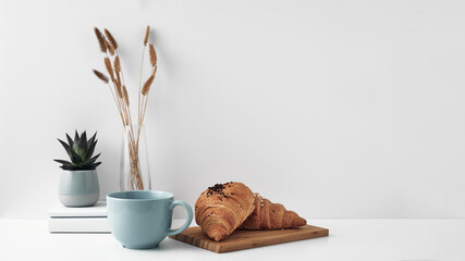 Croissant and a mug on a wooden stand, a vase and a pot with a plant on the books, white background. Eco-friendly materials in interior decor, minimalism. Copy space, mock up