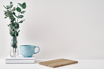 A mug, a stack of books, a wooden board and a transparent vase with eucalyptus branches....