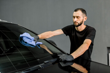 Fototapeta na wymiar Car wash and cleaning at professional auto service station. Shot of handsome bearded young male worker in black t-shirt and gloves, cleaning car windscreen with blue microfiber cloth