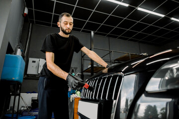 Fototapeta na wymiar Detailing manual cleaning with soap at car wash. Young male worker in rubber gloves washes car radiator grille of luxury black car with special brush and soap. Car detailing wash.