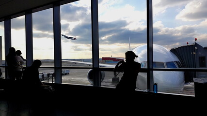 Menschen am Flughafen vor Glasscheibe mit zwei Flugzeugen im HIntergrund davon einem startenden...