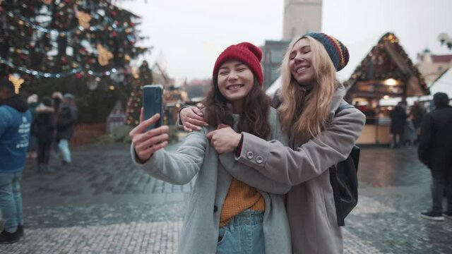 Portrait of two girls making selfie in center of square in european town. Girls taking photo in heart of christmas fare. Background of christmas tree and decorations. Concept of travel, gadgets.