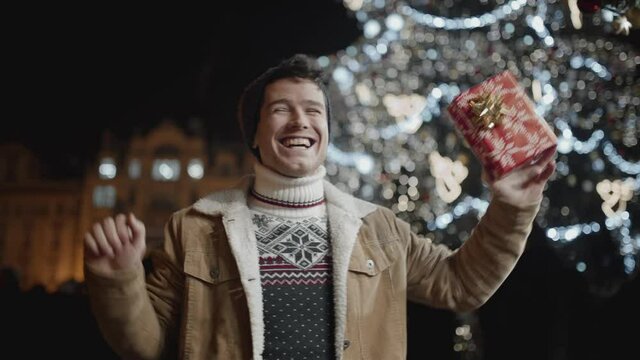 Portrait of happy guy dancing in a middle of a square during holiday season at night. Teenager smiling, celebrating in front of tall christmas tree.