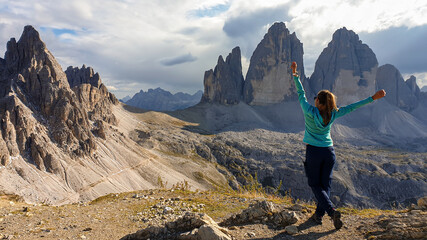 A woman in hiking outfit standing with her arms wide open and enjoying the view on the famous Tre Cime di Lavaredo (Drei Zinnen), mountains in Italian Dolomites. Desolated and raw landscape. Freedom