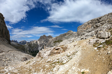 A panoramic view on Dolomites in Italy. There are sharp and steep mountain slopes around. Lots  of lose stones and pebbles. The sky is full of soft clouds. Raw landscape. Serenity and calmness