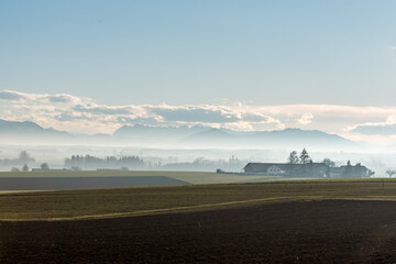 Alpen Panorama im Chiemgau  in Bayern bei Föhn  im Winter mit  leichtem Nebel , blauem Himmel  Wolken und Sonnenschein mit Häuser, Bäumen und Bergen