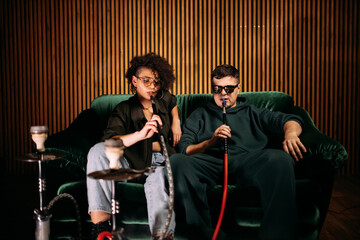 Man and woman chilling on sofa and smoking hookah kalian. Couple relaxing and smoking in studio