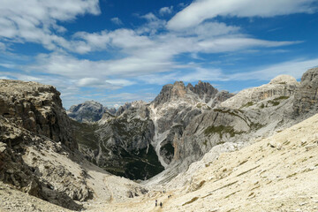A panoramic view on Dolomites in Italy. There are sharp and steep mountain slopes around. Lots  of lose stones and pebbles. The sky is full of soft clouds. Raw landscape. Serenity and calmness