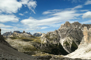 A panoramic view on Dolomites in Italy. There are sharp and steep mountain slopes around. Lots  of lose stones and pebbles. The sky is full of soft clouds. Raw landscape. Serenity and calmness