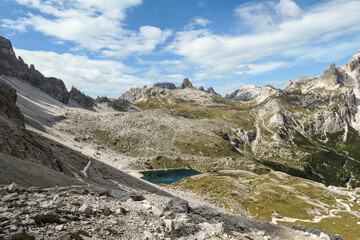Fototapeta na wymiar A panoramic view on Dolomites in Italy. There are sharp and steep mountain slopes around. At the bottom of a small valley there is a small navy blue lake. The sky is full of soft clouds. Raw landscape