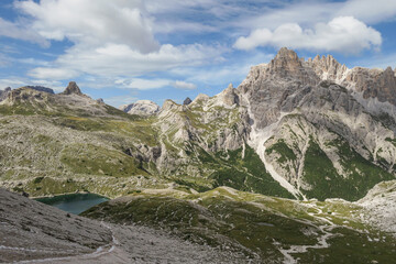 Fototapeta na wymiar A panoramic view on Dolomites in Italy. There are sharp and steep mountain slopes around. At the bottom of a small valley there is a small navy blue lake. The sky is full of soft clouds. Raw landscape
