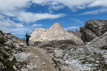 Fototapeta na wymiar A woman with backpack and sticks hiking on a narrow path in Italian Dolomites. There are sharp and steep mountains. At the bottom of a small valley there is a small, navy blue lake. Raw landscape.
