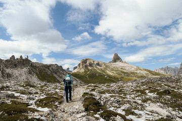 Fototapeta premium A woman with backpack and sticks hiking on a narrow path in Italian Dolomites. There are sharp and steep mountains. At the bottom of a small valley there is a small, navy blue lake. Raw landscape.