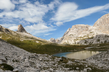 Obraz premium A small, navy blue lake at the bottom of the valley in Italian Alps. The lake is surrounded by high and steep peaks. The slopes are lush green. The sky is full of soft clouds. Raw landscape. Remedy
