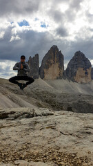 Naklejka na ściany i meble Man in hiking outfit jumping in front of the famous Tre Cime di Lavaredo (Drei Zinnen), mountains in Italian Dolomites. Desolated and raw landscape, full of lose stones. Overcast. Achievement and fun