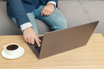Unrecognized man, businessman or freelancer sitting on sofa and working at laptop on table with coffee cup, work from home