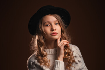 Charming blonde teen girl in black hat posing in studio on brown background