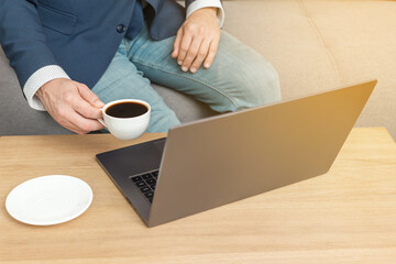Unrecognized man, businessman or freelancer sitting on sofa, drinks coffee and working at laptop, work from home