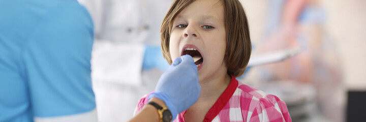 Doctor in blue suit look at child throat with wooden stick. Child in plaid shirt sit at doctor...