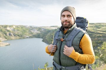 Naklejka premium Happy young male backpacker standing against river surrounded by mountains