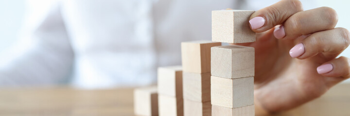 Woman build tower from wooden cubes on table close-up. Career growth up stairs. Female hand put...