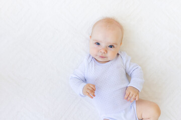 portrait of a small baby girl 6 months old in a white bodysuit lying on her back on a white bed, space for text
