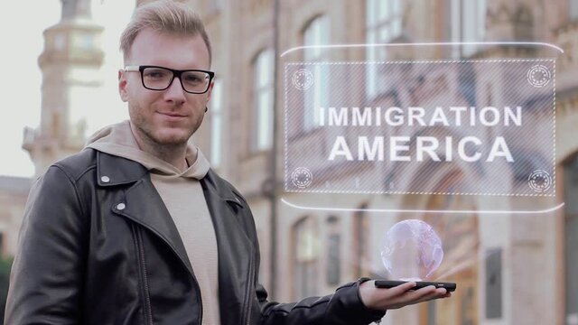 Smart young man with glasses shows a conceptual hologram Immigration America. Student in casual clothes with future technology mobile screen on university background