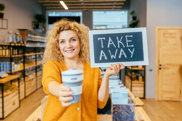 Young blond curly woman holding sign board with Take away text and silicone collapsible cup on...