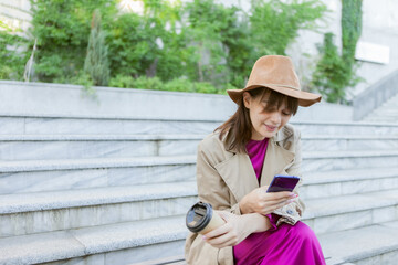 Obraz premium Young fashion woman dressed in autumn clothes sits on bench and uses a smartphone outdoors